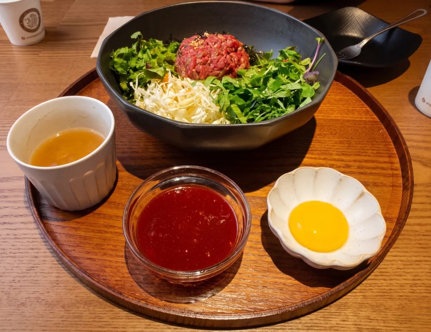 Wooden tray with a bowl of greens and raw beef tartare, a cup of broth, dipping sauce and a raw egg yolk
