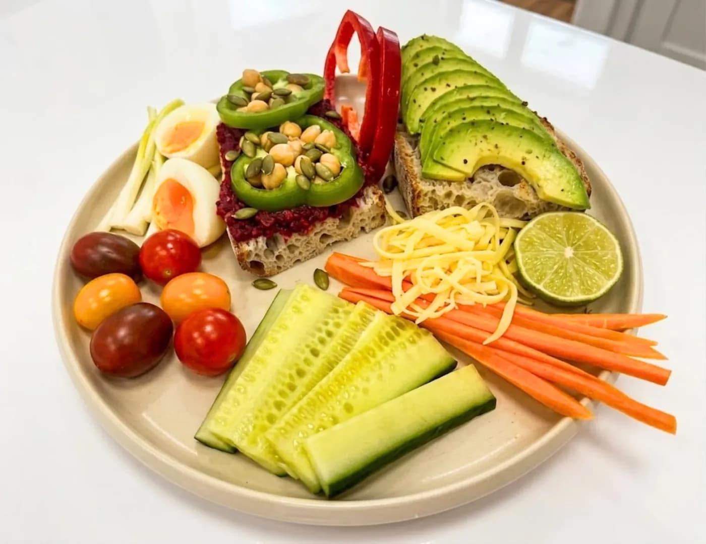 Plate of avocado and beetroot-hummus toast with a boiled egg, cheese, carrot and cucumber sticks, cherry tomatoes and lime