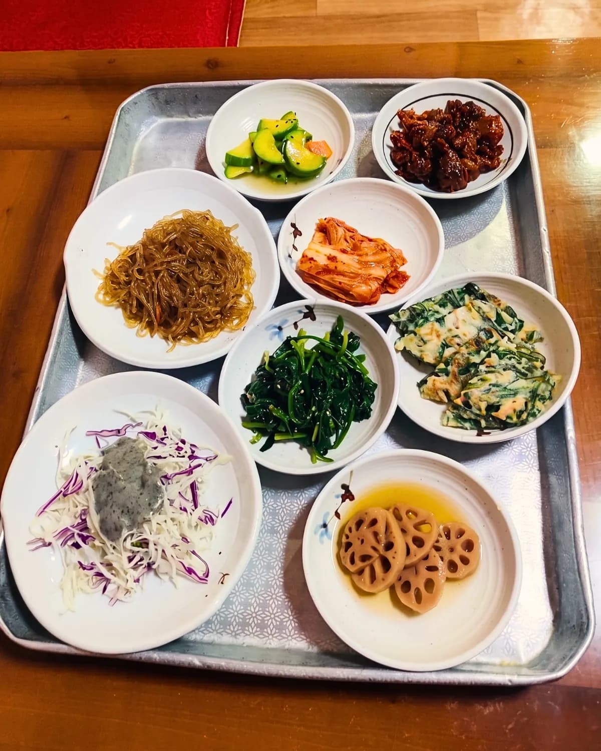 Tray of small bowls of assorted Korean side dishes including kimchi, glass noodles and lotus root