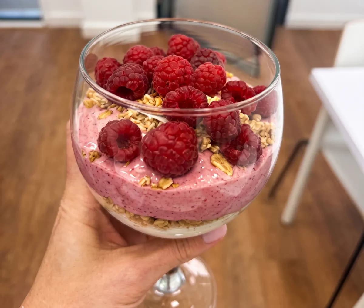 Hand holding a glass of raspberry yoghurt parfait layered with granola